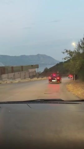 The video is taken from the perspective of a person inside a car, looking out through the windshield. It appears to be dusk or early morning, with a pale sky and a distant mountain range. A road curves to the right, with several cars driving ahead, their brake lights illuminated. The car the video is taken from is also moving. On the left side of the road, there is a building with a fence. On the right side, there are trees and a street light. The overall scene suggests a scenic drive along a coastal or hilly area as daylight fades.