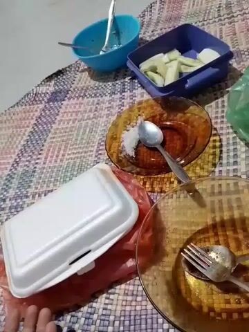 The video shows a close-up, high-angle view of food and dishes on a woven mat. In the foreground, a white styrofoam container filled with meatballs in a reddish sauce is open. To the right, a brown glass plate holds a spoon and a pile of white granular substance, possibly salt. Another brown glass plate with a fork and spoon rests below it. In the background, a blue bowl contains remnants of food and a fork, and a blue rectangular container holds sliced green vegetables. A red plastic bag is partially visible beneath the styrofoam container. The lighting suggests an indoor setting, possibly in the evening.