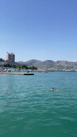 This media file captures a bright, sunny day at a coastal location, likely in Sadovyi, Russia, given the provided context. The scene is dominated by a vast expanse of clear, turquoise water, gently rippling under a bright blue sky. In the foreground, a person is swimming, their head and shoulders visible above the water, enjoying the refreshing sea.

Further out in the water, other individuals can be seen engaged in various activities. Some appear to be swimming or wading closer to shore, while others might be on floatation devices, indicated by distant, small shapes.

Along the coastline, a town or city stretches out, characterized by buildings of varying heights, including a prominent multi-story structure that appears to be a hotel or apartment complex. A pier extends into the water, with people milling about on it, suggesting it's a recreational area or a docking point. Beyond the town, a range of rugged, layered mountains forms the backdrop, their slopes appearing somewhat barren and possibly quarried in parts, with industrial structures and cranes visible on the lower slopes and near the coast. These industrial elements, along with the presence of a harbor or port, suggest a working coastal town.

The overall atmosphere is one of a warm, pleasant summer day. The clear sky and calm water indicate favorable weather conditions for outdoor activities. The presence of swimmers and people on the pier points to a popular tourist or recreational spot. The bright sunlight suggests it is midday or early afternoon. No visible text is present in the media.