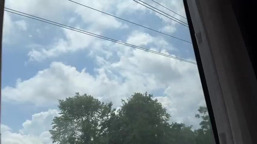 A view from inside a train window shows a bright blue sky filled with puffy white clouds. Overhead, power lines for the train stretch across the frame. On the ground below, green trees are visible. The camera pans slightly to the right, revealing more of the overhead power line infrastructure, including poles and complex wiring. Further along, a metal structure supporting the power lines is seen. The scene captures the experience of traveling by train on a clear day, with the sky and surrounding landscape passing by the window.