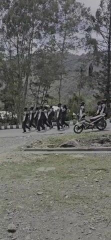 A group of young people in uniform are marching in formation on a paved road. They are wearing dark uniforms with white accents and appear to be performing some kind of drill or parade. Some of them are carrying what look like instruments, suggesting they might be a marching band. The background shows a natural environment with trees and a mountain in the distance. A motorcycle is parked on the side of the road. The lighting suggests it is daytime.