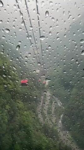 The video shows a view from inside a cable car on a rainy day. Raindrops cover the window, obscuring the view outside. Two other red cable cars can be seen further up the line. Below the cable cars is a hillside covered in lush green trees and a winding road. The sky is overcast and gray.  The overall scene is hazy due to the rain and fog.