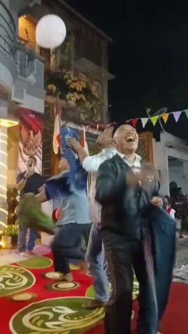 A group of people are gathered outdoors at night, possibly for a celebration. Several people are looking up at the sky, and some are holding fabrics. There are balloons in the air, one white and one red. One person is wearing a dark jacket with a design on the back that says "ZADE". The ground is covered in a patterned carpet. There are lights and some building structures in the background. The overall atmosphere appears to be festive and energetic.