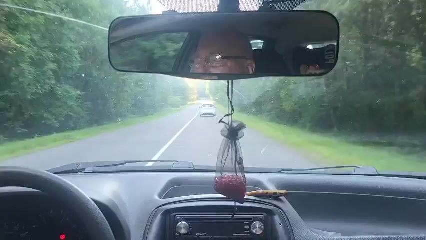 The video shows the interior of a car driving on a paved road. The perspective is from the driver's seat, looking forward through the windshield. The steering wheel is visible on the left. The dashboard has a car stereo system and an air freshener hanging from the rearview mirror. The rearview mirror reflects the face of the driver, who appears to be wearing glasses. Outside the car, trees line both sides of the road, and there is a white car driving ahead. Another white car is visible passing the driver's car on the left. The road has a white line marking the center.