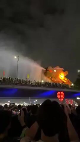 At night, a crowd of people observes a water-spraying dragon sculpture on a bridge. Most of the crowd holds up phones to record the spectacle. The dragon sculpture is lit up with orange lights and is set against the dark sky. The bridge is illuminated with blue lights, and people are standing along its length, some waving flags. A lamppost provides additional lighting on the bridge. The environment appears urban, and the atmosphere is celebratory. The overall scene conveys a sense of awe and entertainment.
