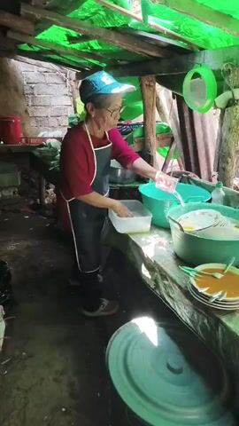 An elderly woman wearing glasses, a red shirt, and a blue baseball cap is washing dishes in an outdoor kitchen. She is standing at a counter covered with a green tarp. In front of her is a white tub filled with water and dishes. To her right are several large green tubs overflowing with dirty dishes, plates, and bowls. The kitchen has a rustic appearance, with a wooden roof covered in green plastic sheeting and corrugated metal walls. The floor is dirt. The overall impression is of a simple, functional outdoor kitchen.
