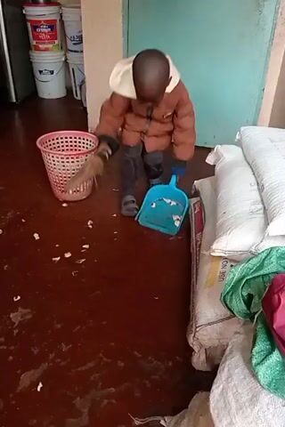 A small child wearing a puffy brown jacket is cleaning a red-brown floor in a room. They are using a small blue dustpan and a broom to sweep up small white pieces. A pink plastic wastebasket sits nearby. Behind the child are several white sacks, partially covered with a green cloth. Behind the wastebasket are several white buckets. A gas cylinder is visible on the left side.  The room has a painted light-blue wall on one side and a partially visible light-peach wall on another.
