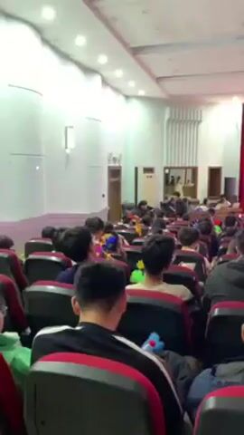The video begins with an audience seated in an auditorium, facing a stage with a large screen. Most attendees are young men. The screen displays an image, and the room lights are dimmed. Some attendees are looking at their phones. The video then shifts focus to a pair of legs in jeans and white sneakers, with the person walking on a speckled carpet. The camera is then briefly pointed under a row of seats, showing a glimpse of a pink object underneath.