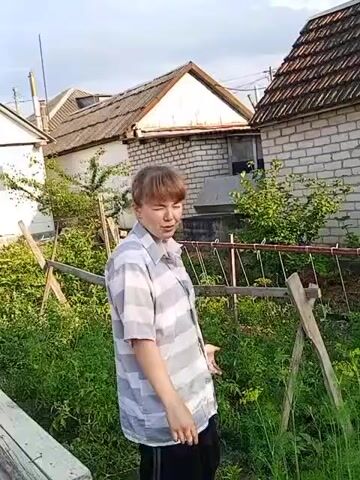 A person with short, reddish-brown hair and wearing a grey and white striped button-down shirt and black pants stands in a garden. The person's left hand is extended and slightly open, as if gesturing. The person's head is turned slightly to their right, and their gaze appears to be directed forward. The background shows two buildings constructed with brick and a tiled roof, and an abundance of green foliage. Wooden stakes and a string or wire are visible in the garden area. The sky above is overcast.