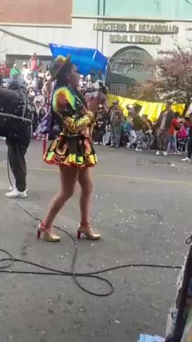 The video shows several women dancing in a parade. The women are wearing colorful, elaborate costumes with short skirts, hats, and gold high heels. They are performing in front of a crowd of spectators. The background features buildings, including one with the words "Ministerio de Desarrollo Rural y Tierras." There are poles and a metal cart in the foreground, partially obstructing the view. A man with a camera is standing to the left of the women. The street is paved and appears to be closed off for the event.