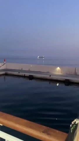 The video begins with a view of a calm body of water, possibly a harbor or bay, under a twilight sky. A distant cargo ship is visible on the horizon, its lights reflecting on the water. A concrete pier extends into the water, with a red navigation buoy on the left and several lights illuminating the walkway. The camera then pans to the right, showing the side of a vessel, likely a ferry or cruise ship, with a wooden handrail in the foreground. The interior of the ship is partially visible, with people walking on a deck. The video ends with a shot of the side of the ship against the water, with more people visible on the deck.