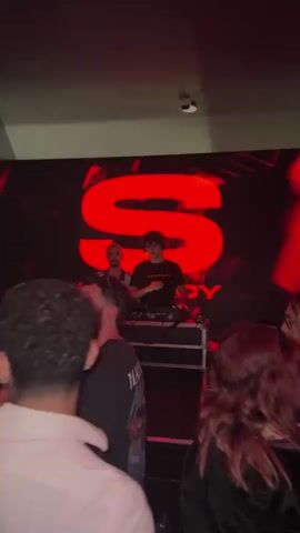This video captures a vibrant nightlife scene inside a club or event space in Turin, Italy. The setting is dimly lit, dominated by a striking red glow emanating from a large LED screen at the back.

At the center, two DJs are actively performing behind a DJ booth, surrounded by various equipment including mixers and turntables. One DJ wears a black t-shirt with "SEXSTASY" printed on it, while the other wears a white and blue patterned top. Drinks are placed on the DJ booth, suggesting a lively atmosphere.

The large screen behind them displays a prominent, stylized red letter "S," with faint text that appears to be "SEXSTASY" visible below it, matching the DJ's attire. The red lighting casts dramatic shadows and highlights across the space.

In the foreground, numerous attendees are visible, enjoying the music and dancing. Their faces are partially obscured by the low light, but their postures and movements suggest an energetic and engaging atmosphere. The crowd consists mainly of young adults, dressed casually for a night out. The overall mood is one of excitement and revelry, characteristic of a club event.