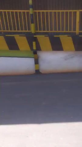 The video shows a stationary camera facing a loading dock area. In the foreground, there is a paved surface with a distinct shadow. Behind the shadow, there is a long, white, cylindrical object, possibly a bumper or dock seal, positioned horizontally. Above this, there is a black and yellow striped barrier. Further back, a yellow metal fence is visible. To the right of the fence, a sign with text and images is mounted on a wall. The wall appears to be made of corrugated metal. The overall environment seems to be an industrial or commercial setting, possibly a warehouse or loading bay.