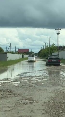 The video shows two cars driving through a flooded street. The street is unpaved and full of puddles and gravel. The water is muddy and brown. On either side of the street are fences, telephone poles, and small trees and bushes. The sky is overcast with dark storm clouds, although there are hints of blue sky visible. A white van drives towards the viewer, while a black sedan travels towards the background, both kicking up water. The license plates on the black car are visible but difficult to read. In the distance, another vehicle can be seen. The video appears to be taken from inside a vehicle moving along the flooded street.
