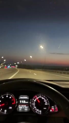 A first-person perspective video shows the interior of a car driving at night. The dashboard is visible, with the speedometer reading 113 km/h at 0:00 and increasing to 114 km/h by 0:02. The sky is dark blue with hints of pink and orange from a sunset, and streetlights line the highway. The road ahead is lit, with other cars visible in the distance. The steering wheel is visible in the foreground.
