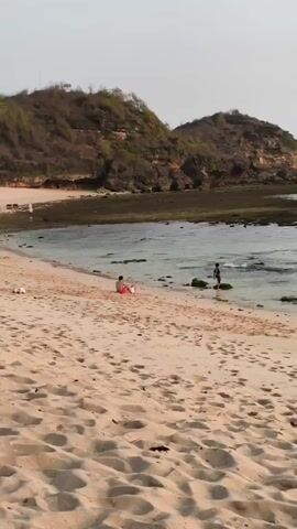 The video shows a sandy beach with two people. One person is sitting on the sand in an orange and white outfit, facing the ocean. Another person, wearing dark clothing, is standing in shallow water near some rocks, facing away from the camera. Behind the beach are two hills covered in dry vegetation, and the water is calm with small waves. The sky is overcast and light-colored.