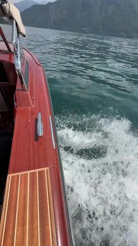 The video shows the side of a wooden boat moving through water, creating a wake. The boat has a polished wooden exterior and chrome accents. In the background, a body of water stretches to a distant shore with hills and mountains. The sky appears to be sunny.