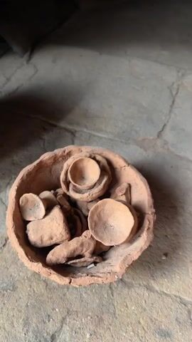 The video shows a display of handmade terracotta pots and other items on a stone shelf within a stone alcove, which appears to be part of a workshop or storage area. The pots are various sizes and shapes, including a large pot with a lid, cylinders, vases, and two cone-shaped items. In the center, at the bottom, is a bowl containing miniature clay pots. In the bottom right corner is an orange soda bottle. The lighting is dim inside the alcove, highlighting the texture of the stone and clay.
