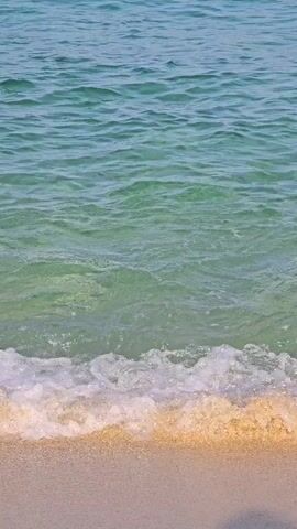 A video shows the gentle lapping of clear turquoise water onto a sandy beach. The water's surface is slightly rippled, and sunlight refracts through it, illuminating the sand beneath the shallow waves. The scene is tranquil and focuses on the natural beauty of the ocean meeting the shore.