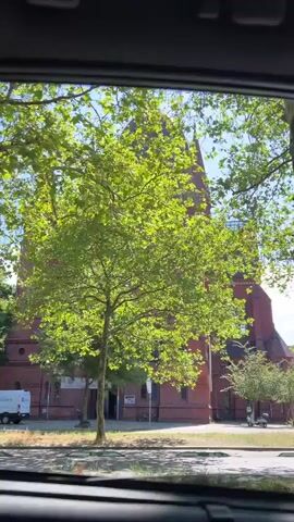 The video, filmed from inside a vehicle, captures a large, red brick church with a distinctive domed tower and pointed spires. The camera pans slowly to the right, revealing more of the church's facade, including arched windows and a large circular window above the entrance. Green trees with full foliage are visible in the foreground and surrounding the church. The sky is blue with scattered white clouds. A few people are seen walking on a sidewalk and crossing a street in front of the church. Traffic lights and street signs are also present. The scene appears to be a sunny day in an urban or suburban environment.