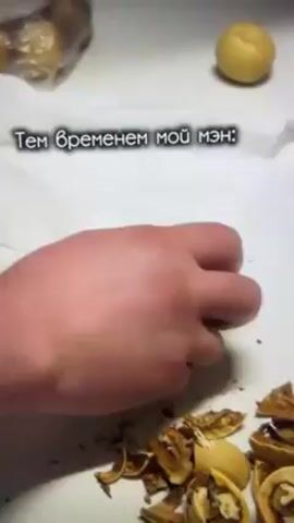 A pair of hands rolls a yellow ball of dough in a white square of paper towel.  The man places nut shells aside, and another ball of dough sits in the background. The man's hands are red. A bag of dough is in the background. The text "Тем временем мой мэн:" is on screen.