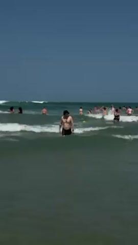 The video shows a sunny beach scene with numerous people in the ocean. Waves are visible. One man wearing shorts and a cap stands in the water, facing towards the camera. Other people are swimming, standing, or interacting with the water in the background. The sky is clear and blue.