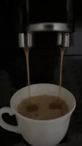 In a close-up shot, an espresso machine dispenses coffee into a white ceramic mug. The dark brown coffee pours from two nozzles into the cup, which is already partially filled with a light-colored liquid, presumably milk or creamer. This creates a dark splash in the light-colored liquid. The machine is mostly black with silver accents, and the mug rests on a metallic drip tray with circular indentations. The lighting is somewhat dim, emphasizing the contrast between the dark coffee and the white mug. The camera remains static throughout the brief video.