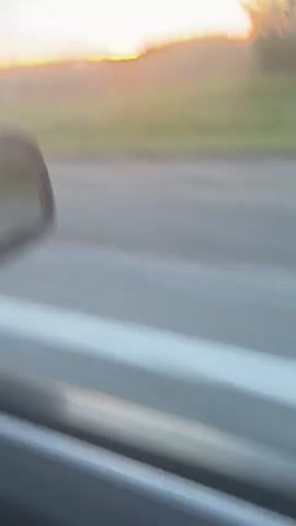 The video is a blurry, handheld shot from inside a moving vehicle, capturing a sunset over a rural landscape. The side-view mirror of the car is partially visible on the left side of the frame. The sky displays hues of orange, yellow, and blue with scattered clouds. A line of trees marks the horizon, and green fields are visible in the middle ground. The foreground shows a gray road, indicating the vehicle is in motion. The overall impression is one of a fleeting view of a natural scene during twilight.