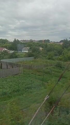 The video shows a view from a moving vehicle, likely a train, looking out at a rural landscape under a cloudy sky. The scene features several houses with wooden fences and gardens. In the foreground, there is a field of green plants, possibly vegetables, bordered by a wooden fence. Further back, there are more houses, some with colorful roofs, nestled among trees. A church steeple can be seen in the distance. The overall impression is one of a quiet, rural village or countryside.