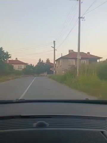 The video shows a view from the inside of a car driving down a paved road in what appears to be a village. On the right side of the road are houses and overgrown vegetation. There are power lines overhead. In the distance, there is a stop sign and a barrier across the road. The sky is a pale blue, suggesting it is either early morning or late evening.
