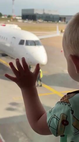 A young child's left hand is held up to a window, with fingers spread. The child's arm and shoulder are visible in the foreground, wearing a light green shirt with a pattern of cartoon dinosaurs and skateboards. Outside the window, a white airplane is parked on the tarmac. Yellow lines are painted on the tarmac. In the background, other airplanes and airport buildings are visible. Two airport workers in yellow vests are near the parked airplane. The child's face is not fully visible, but their mouth is slightly pursed. The child appears to be looking at the airplane.