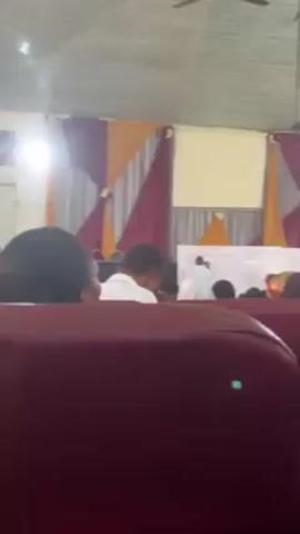 The video shows the back view of an audience in a room. A person is standing at the front of the room near a whiteboard, writing with a marker. Behind them are maroon, silver, and gold decorations hanging on the wall. A projector screen is also hanging on the wall. The audience is sitting in red upholstered chairs. Some people are facing forward, while others are looking down or to the side. The ceiling of the room is light green and has several light fixtures and ceiling fans. The overall lighting in the room appears bright.
