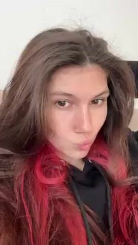 The video shows a woman with long, brown hair that has sections dyed bright pink around her face. She is wearing a black hoodie and has a nose piercing. She is lying down, looking directly at the camera, and touches her hair several times. There is no other significant detail.