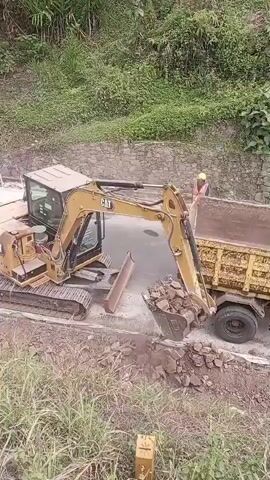 An excavator is loading dirt and rocks into the bed of a yellow dump truck. A person wearing a hard hat and orange safety vest stands near the side of the dump truck, supervising the operation. The scene appears to be on a hillside with lush green vegetation. The ground is uneven and rocky.