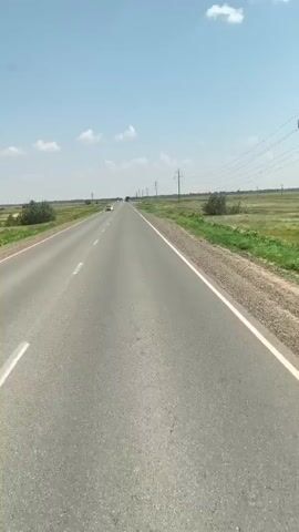 The video begins with a view of an empty highway stretching into the distance under a clear blue sky with scattered clouds. The road is asphalt with white lane markings. The surrounding landscape is flat and covered with dry grass and some green vegetation. A few trees are visible along the roadside. Power lines run parallel to the road in the distance. A white car is seen in the left lane, driving away from the camera. The perspective then shifts to the interior of a vehicle, showing the dashboard and steering wheel. The speedometer needle is pointing slightly above 70, indicating a speed of approximately 77 km/h. The odometer reads 155362. Other gauges and digital displays are also visible. The video then returns to the highway view, showing the road ahead, power lines, and the vast, flat landscape. The sun appears to be high in the sky, suggesting it is daytime. The overall impression is of a journey on a long, straight road through a rural or semi-arid environment.