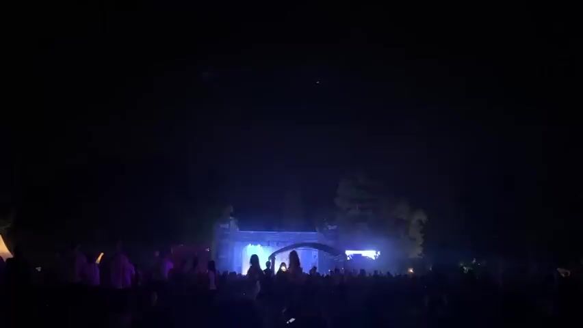 This video captures a nighttime outdoor event, likely a concert or festival, with a crowd gathered in front of a stage. The stage features illuminated screens displaying vibrant colors, primarily purples and blues, which are also reflected in the lighting effects. Smoke or haze is visible around the stage, adding to the atmosphere. The audience members are mostly silhouetted against the stage lights, with some holding up phones or cameras. The overall environment is dark, emphasizing the illuminated stage and the crowd's presence.
