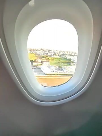 The video is a view from an airplane window looking out at a city. The plane appears to be ascending or descending as the landscape below is visible and detailed. The scene outside the window is a mix of urban development and natural landscape. There are buildings, roads, and what appear to be industrial facilities. In the foreground, there is a large body of brown water or marshland. The sky is clear with a few wispy clouds, and the lighting suggests it is either early morning or late afternoon. The camera is stationary, focused through the airplane window.