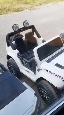 This is a first-person perspective video looking through a window. In the foreground, there is a black and silver object, possibly a car, partially visible. To the right of it is a white toy Jeep Rubicon. The Jeep has black seats, a black roll bar, two large headlights on the roll bar, and a spare tire mounted on the back. The word "Jeep" is visible on the side and front of the vehicle, and "RUBICON" is written on the hood. The Jeep has large, rugged tires. Behind the white Jeep, another toy vehicle, dark gray or black, is partially visible. The ground outside the window is paved with light-colored rectangular stones, and a grassy area is visible in the background. The lighting suggests it is daytime.