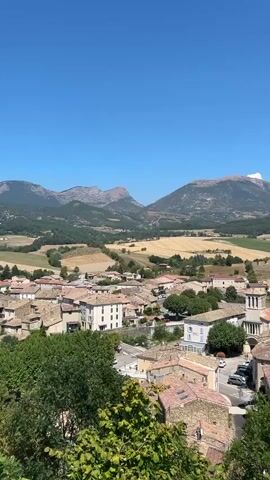 This is a vertical, high-angle, real-time video of a small village nestled in a valley with mountains in the background. The sky is clear and blue. The village has red-tiled roofs and stone buildings, with a church tower prominently visible. Fields of crops, possibly wheat, are scattered throughout the valley. Lush green trees in the foreground partially obscure the view. The timecode indicates the video starts at 00:00:00 and ends at 00:00:05.