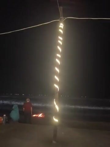 A group of people are gathered on a beach at night. The scene is illuminated by string lights wrapped around a pole in the foreground and two hanging lanterns in the background. A bonfire is visible in the sand. Several individuals are sitting or standing near the bonfire, while others are observing the ocean. The waves are gently crashing on the shore. There are bags and other belongings scattered on the sand. The overall atmosphere appears relaxed and casual.