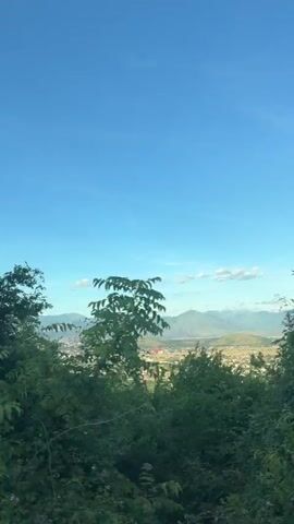 A video taken from a moving vehicle shows a panoramic view of a valley below a mountain range. The sky is a clear blue with a few scattered clouds. Dense green foliage fills the foreground of the shot, obscuring parts of the view. The valley contains a town with buildings, houses, and roads. The video is slightly blurry due to movement.
