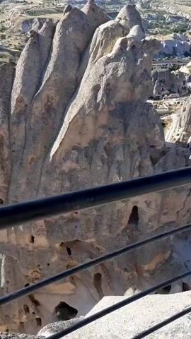 A drone flies over a rock formation with many caves carved into it. The rock is light brown and has many vertical ridges. The caves are dark holes scattered across the rock face. In the background, there is a vast landscape with more rock formations and some vegetation. The sky is blue and clear.