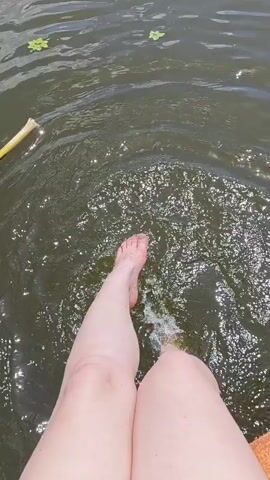 The video shows a person's legs and feet submerged in dark, murky water. The person is kicking their feet, causing ripples and splashing. There are some green lily pads floating on the surface of the water, and a yellow stick is visible in the upper left corner. The lighting suggests it is daytime.