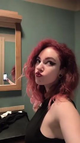 A woman with red wavy hair poses for a selfie video in a bathroom. She wears a black sleeveless top and a decorative necklace. Her makeup includes dark eyeliner and lipstick. The bathroom has green walls and a wooden-framed mirror. The woman initially pouts her lips, then flicks her hair while looking at the camera.