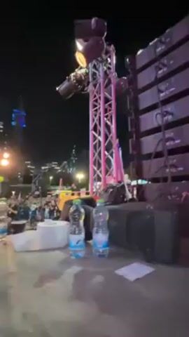 The video shows an outdoor nighttime event with a crowd gathered in front of a stage. Stage lighting is visible along with large sound speakers. There are people holding yellow balloons. The crowd is looking at the stage. Two water bottles are on a platform near the camera.