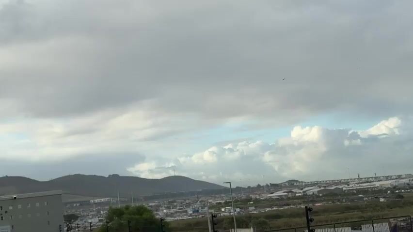 The video shows a daytime shot of a city landscape under a partly cloudy sky. In the foreground, an electric fence runs horizontally across the frame. In the distance, there are buildings and industrial structures, including what appears to be a factory with smokestacks. Behind the city, a mountain range is visible, with Table Mountain dominating the backdrop. The clouds are a mix of gray and white, with patches of blue sky peeking through. The overall scene appears to be a somewhat bleak and overcast day in an urban or industrial area.