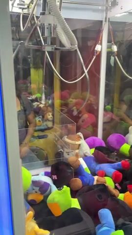 A person is standing in front of a claw machine filled with stuffed animals. The claw is lowered to grab a brown plush item. The inside of the machine is brightly lit. Reflections of the environment and the person can be seen on the glass of the machine. The claw moves around and attempts to grab an item.