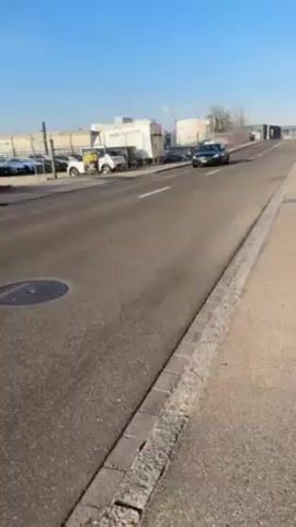 The video depicts a road on a bright, sunny day. A black sedan speeds by on the road. There are several buildings and cars parked alongside the road on the left side. A sidewalk runs along the right side of the road, with a curb separating the road from the sidewalk. A manhole cover is visible on the road. The sky is clear and blue. There are trees visible in the background.