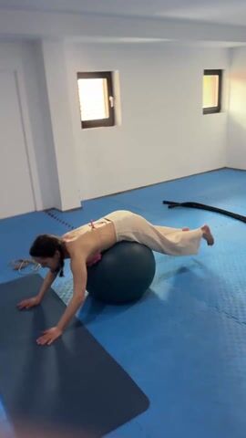 A woman with long dark hair is performing a backbend exercise using an exercise ball and a yoga mat. She is wearing light-colored pants and a pink bikini top. The exercise ball is dark green, and the yoga mat is dark blue. The floor is covered in blue interlocking mats. The room has white walls and a small window. She rests her lower torso on the ball while keeping her arms extended on the mat for support, and she extends her legs upward toward the ceiling.