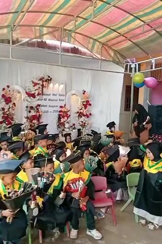 The image captures a graduation ceremony for young children at an educational institution in Keudekrueng, Indonesia. The scene is set under a colorful striped canopy, suggesting an outdoor or semi-outdoor venue. In the background, a white backdrop is decorated with floral arrangements and a sign that reads "Selamat Hari Ceria Anak KB Khairul Amal Tahun Pelajaran 2025-2026," indicating a celebratory event for the "Khairul Amal" kindergarten during the 2025-2026 academic year.

Dozens of children, dressed in black graduation gowns and caps with yellow trim, are seated in rows of colorful chairs. Many of the children are holding bouquets of flowers or other small gifts, likely presented to them as part of the ceremony. The children appear to be excited and engaged, with some looking towards the front, possibly at speakers or performers.

Adults, also wearing hijabs and modest attire, are present, some standing and observing the children, while others appear to be involved in organizing or congratulating the graduates. Two balloons, one yellow and one pink, are visible near the top right of the frame, adding to the festive atmosphere. The overall mood seems joyful and proud, celebrating the children's academic achievements. The lighting suggests it's daytime, likely under warm weather conditions typical for Indonesia.